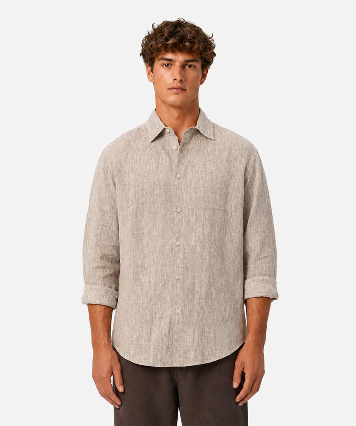 A man wearing a light beige linen button-up shirt with rolled-up sleeves and dark brown trousers, standing against a solid light gray background.