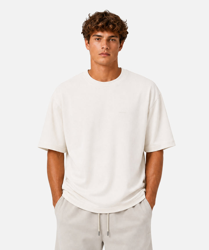 A man wearing a short-sleeve crewneck t-shirt in off-white, paired with light beige drawstring pants, standing against a solid light gray background.