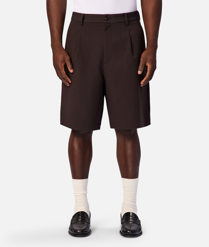 A model wearing dark brown pleated shorts, white crew socks, and black loafers, shown from the waist down against a light gray studio background.