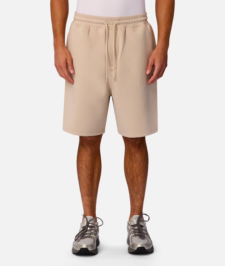 A person wearing light beige drawstring shorts, shown from the waist down against a clean light gray studio background. The shorts have an elastic waistband and a matching drawstring, paired with gray athletic sneakers.