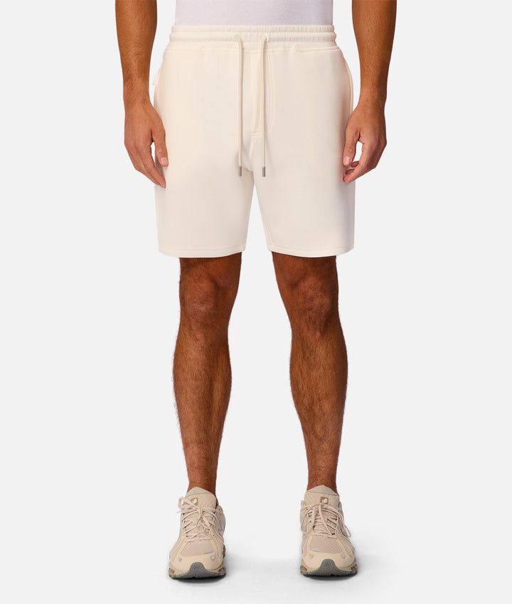 A model from the waist down wearing cream-colored athletic shorts with a matching drawstring, paired with beige sneakers, against a light gray studio background.
