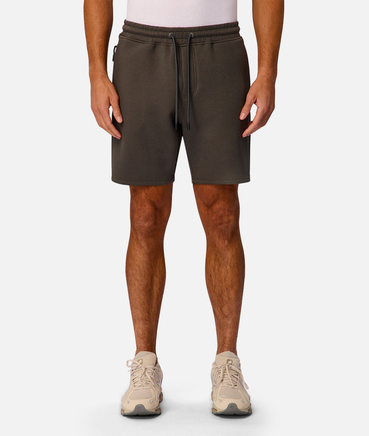 A man wearing dark olive brown drawstring shorts, a light pink t-shirt, and beige sneakers, shown from the waist down against a light gray background.