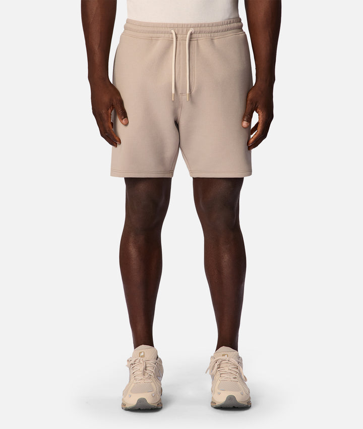 A person wearing light beige drawstring shorts, shown from the waist down against a plain light gray studio background.