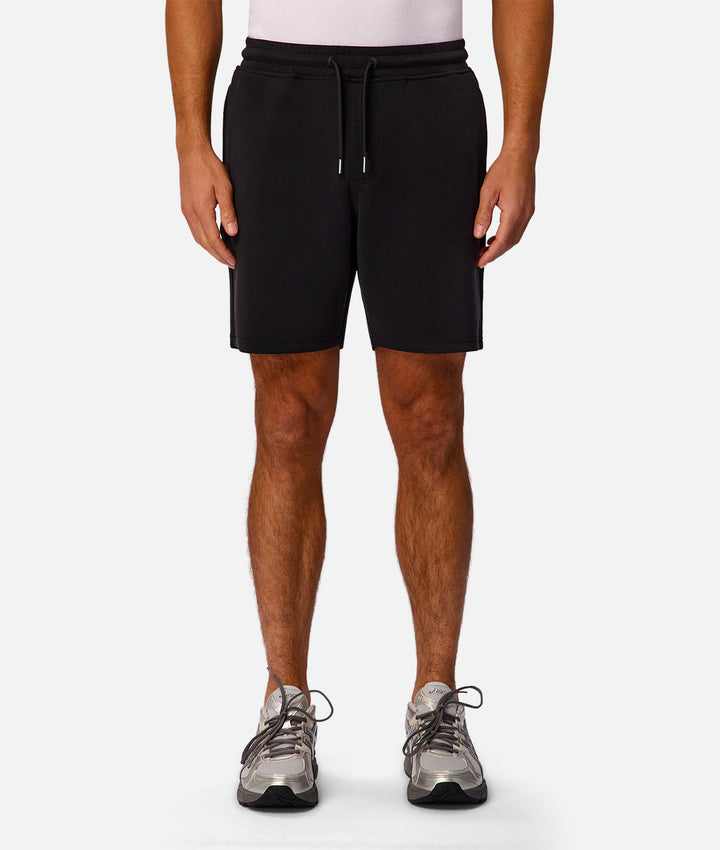 A model from the waist down wearing black athletic shorts with a drawstring waist, paired with gray athletic shoes, against a light gray studio background.