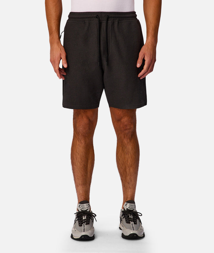 A man wearing black athletic shorts with a drawstring waist, paired with gray and black sneakers, shown from the waist down against a light gray studio background.