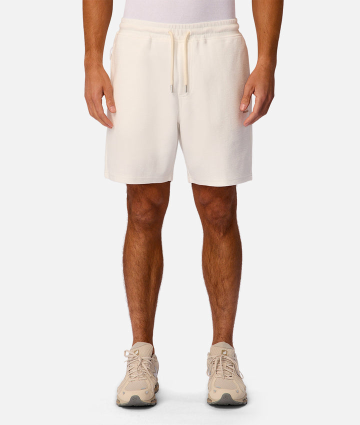 A person wearing off-white drawstring shorts, shown from the waist down against a clean light gray studio background. The shorts have a relaxed fit and a matching drawstring waist.