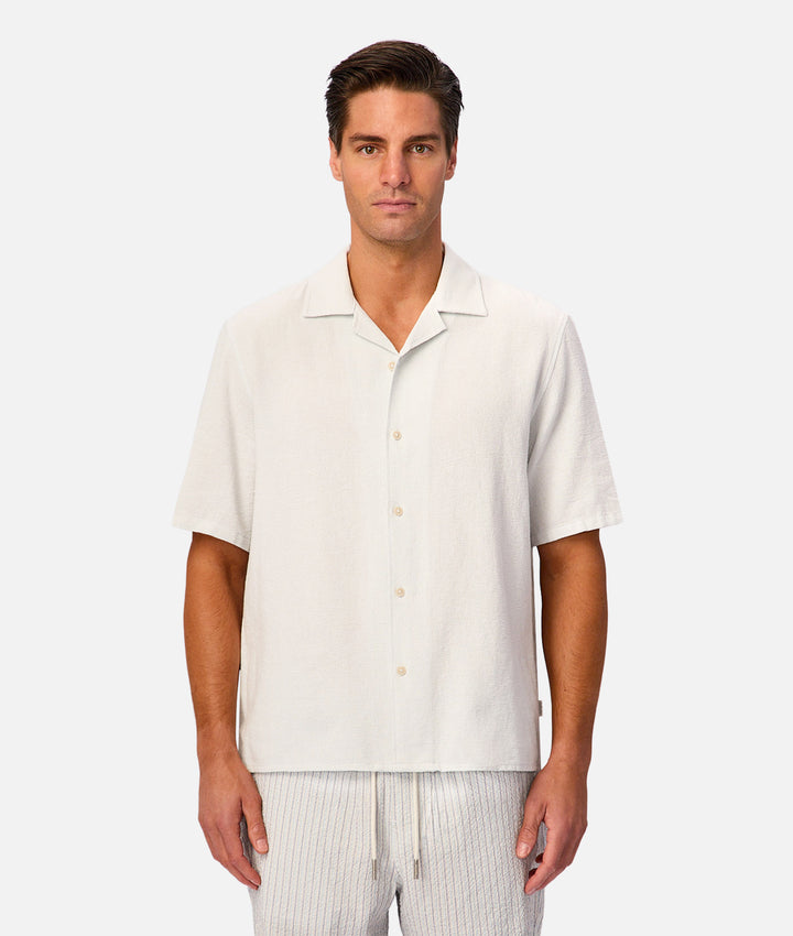 A man wearing a short-sleeve camp collar button-up shirt in off-white linen, paired with light gray striped pants, against a solid light gray background.