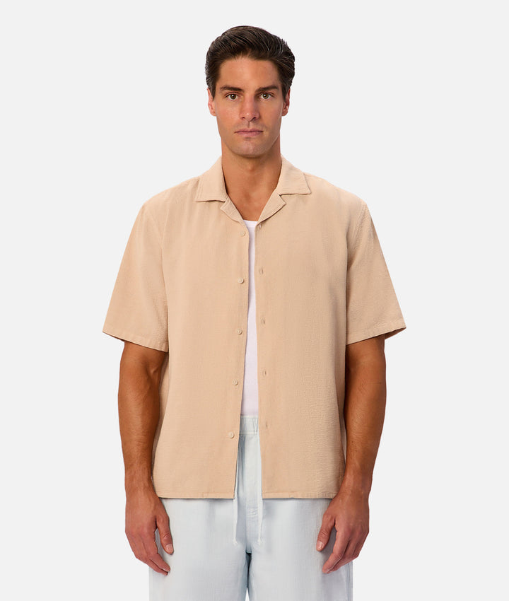 A man wearing a light beige short-sleeve button-up shirt over a white undershirt and white pants, standing against a light gray studio background.
