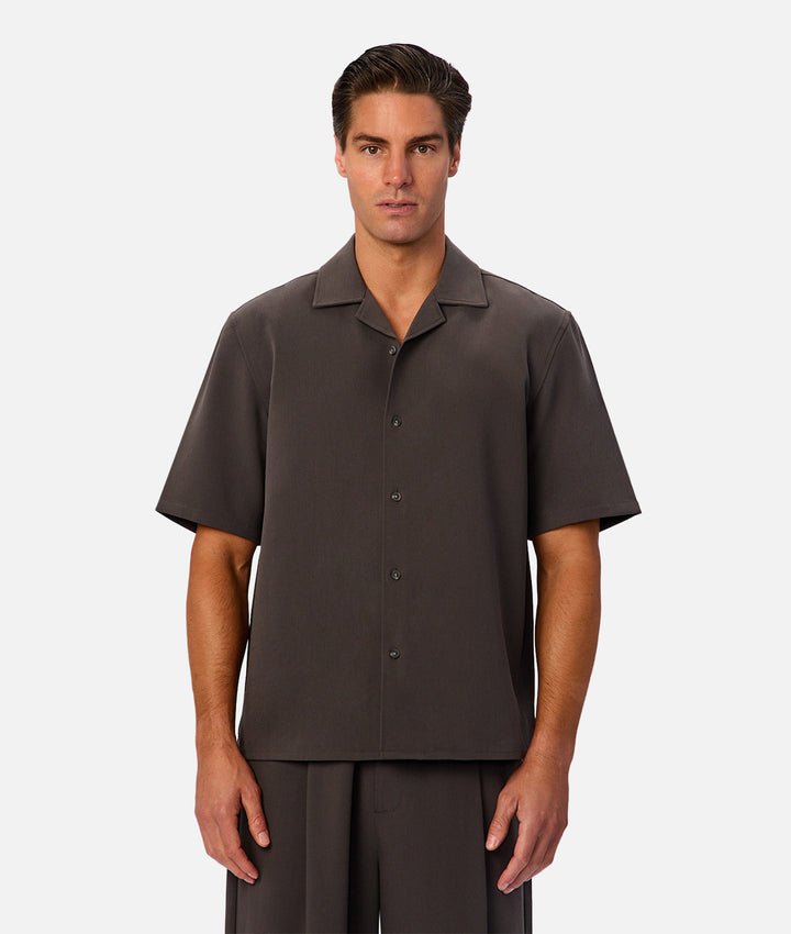 A man modeling a dark brown short-sleeve camp collar button-up shirt and matching trousers against a light gray studio background.