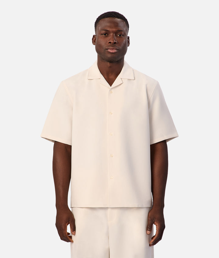 A man wearing a short-sleeve cream-colored camp collar button-up shirt and matching cream trousers, standing against a light gray studio background.