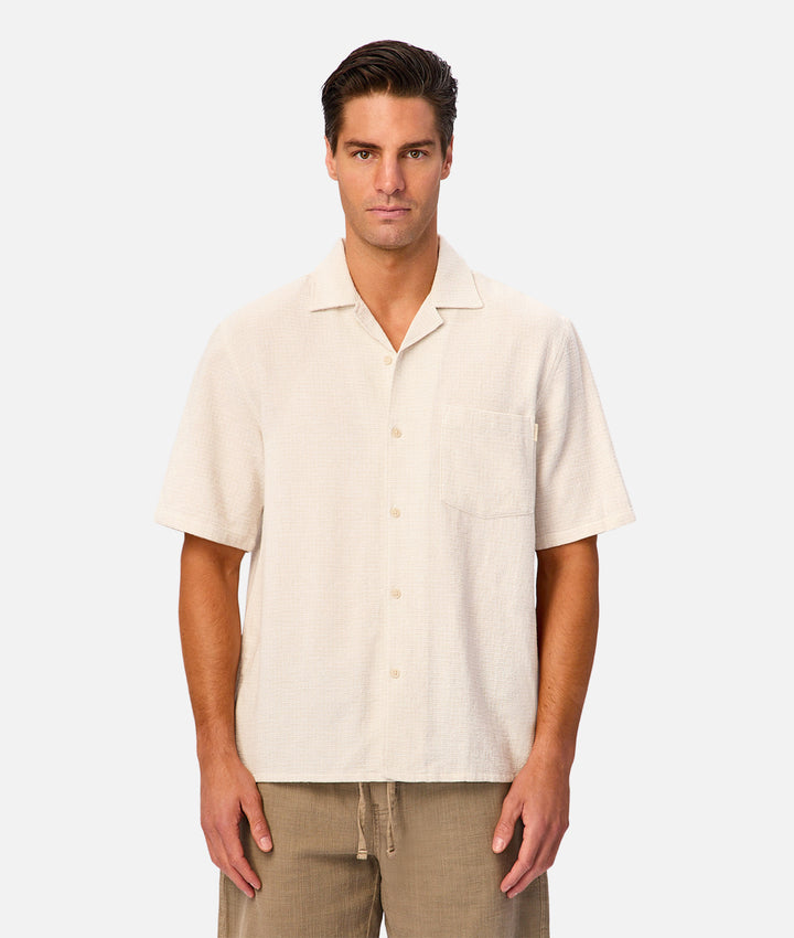 A man wearing a short-sleeve button-up shirt in a light cream or off-white linen-like fabric, paired with khaki pants, standing against a solid light gray background.