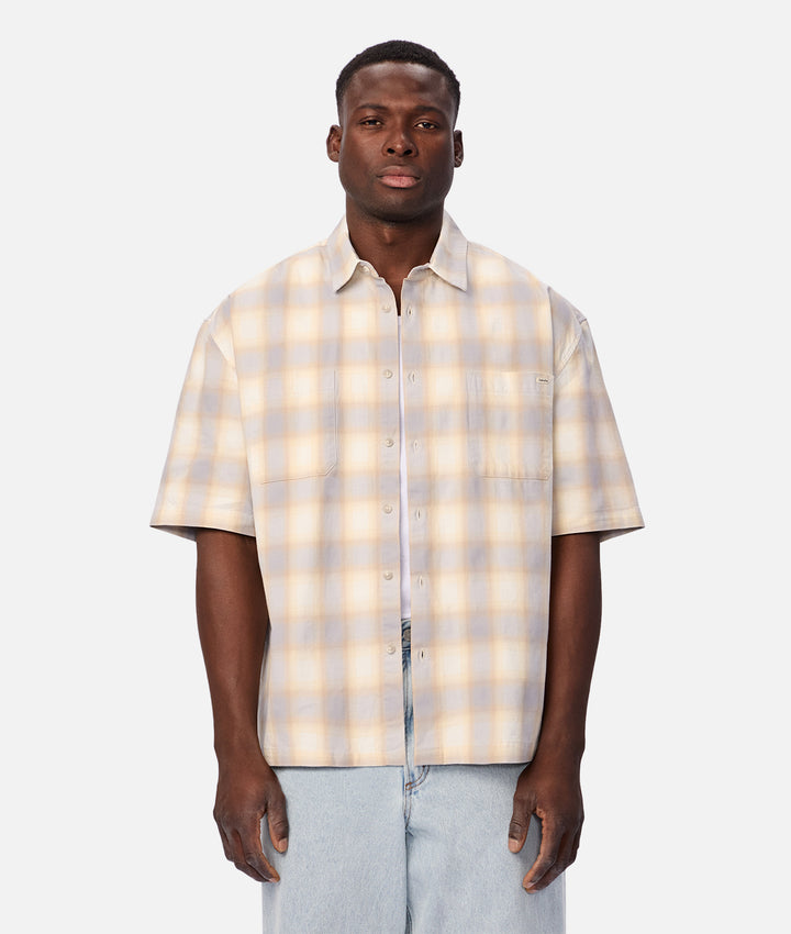 A man wearing a short-sleeve button-up shirt with a light beige and white plaid pattern, paired with light blue jeans, standing against a solid light gray background.