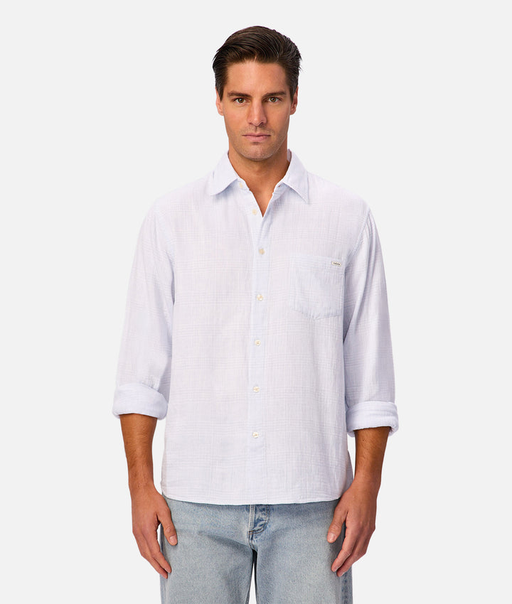 A man wearing a white long-sleeve button-down shirt with rolled-up sleeves and light blue jeans, standing against a light gray studio background.