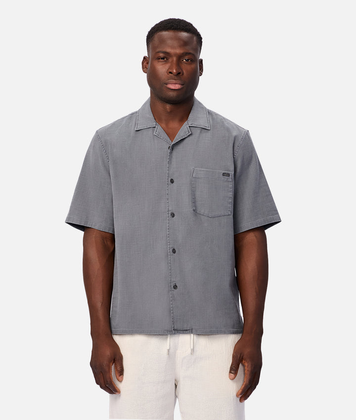 A man wearing a gray short-sleeve button-up camp shirt and white pants, standing against a light gray studio background.