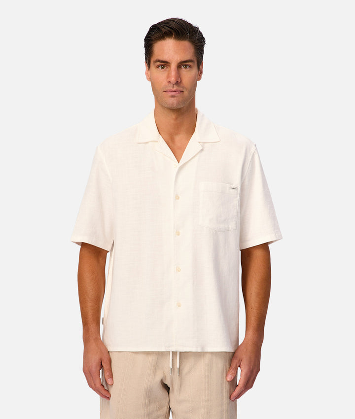 A man wearing a short-sleeve button-up shirt in off-white linen with a chest pocket, paired with light beige pants, against a light gray studio background.