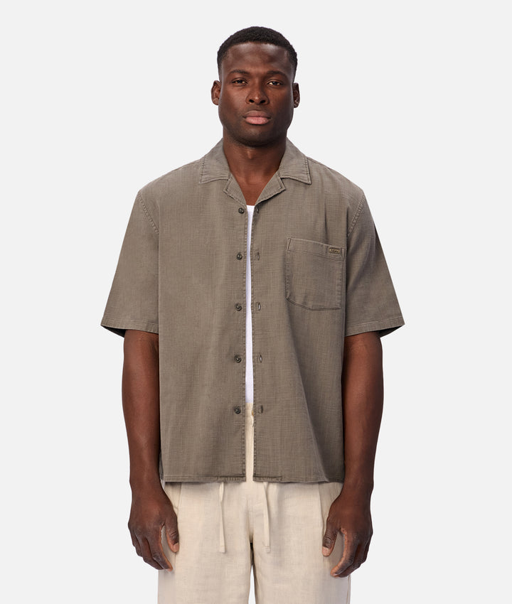 A man wearing a short-sleeve button-up shirt in a textured taupe-brown fabric, paired with light beige drawstring trousers, against a solid light gray background.