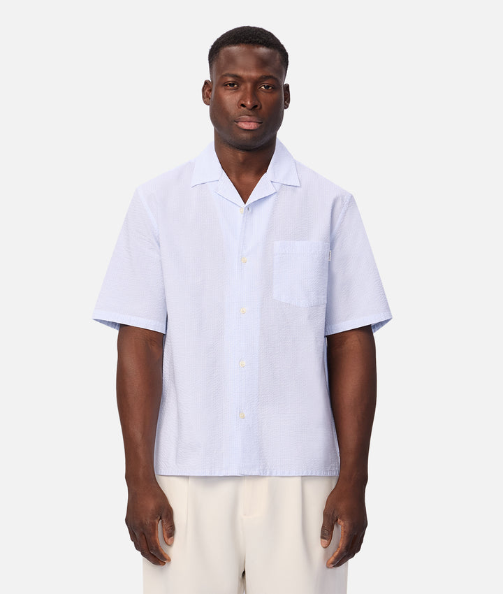 A man wearing a light blue short-sleeve button-up shirt with a chest pocket, paired with cream pants, standing against a light gray studio background.