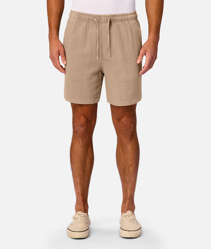 A man wearing beige drawstring shorts, a white t-shirt, and beige sneakers, shown from the waist down against a light gray studio background.