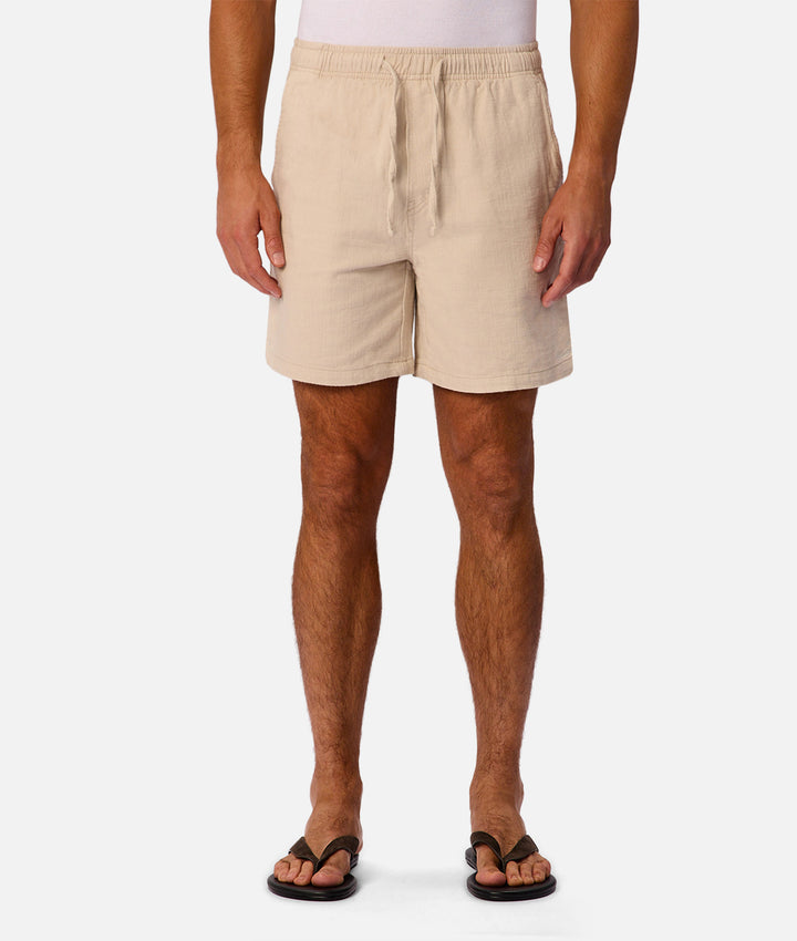 A man wearing beige drawstring shorts and black flip-flops, shown from the waist down against a light gray studio background.