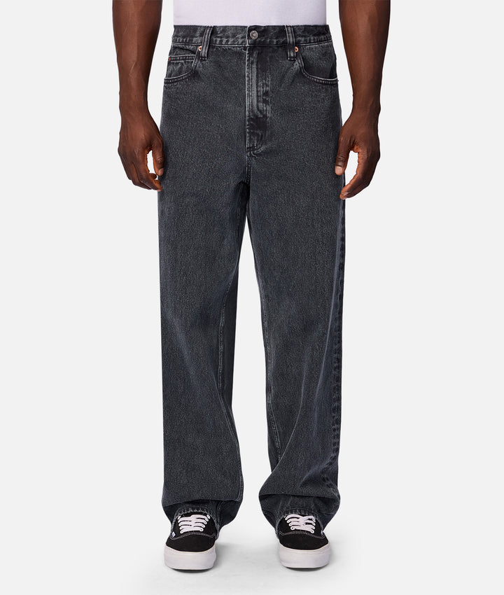 A person wearing loose-fit dark gray washed denim jeans with black low-top sneakers, shown from the waist down against a light gray studio background.