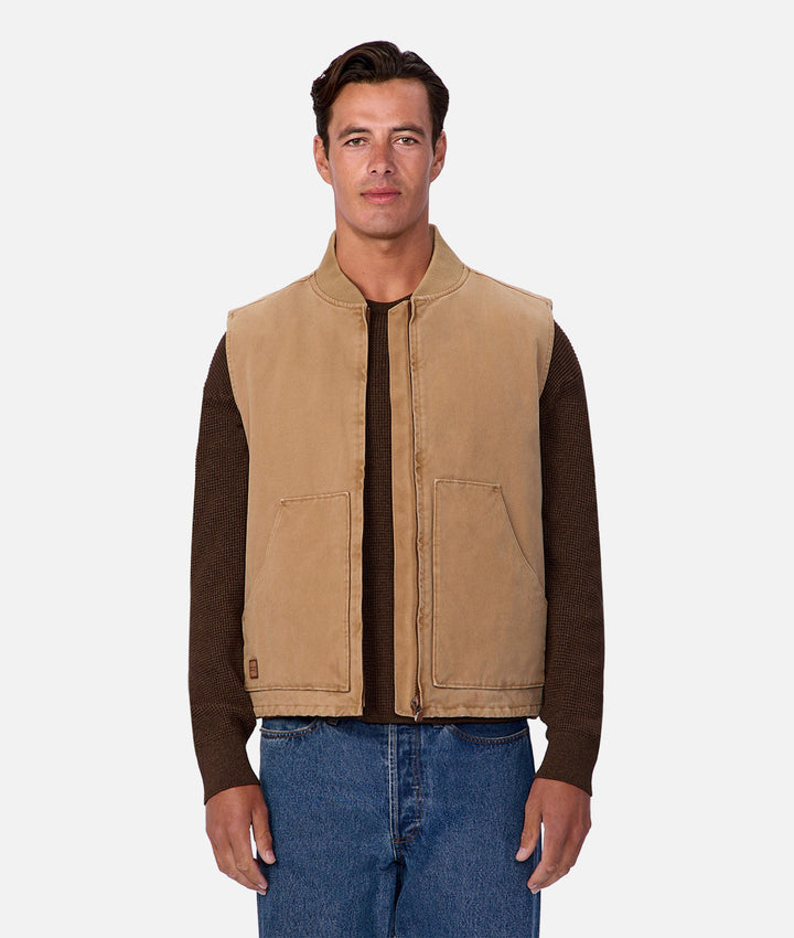 Man wearing a beige vest over a brown shirt with blue jeans on a white background