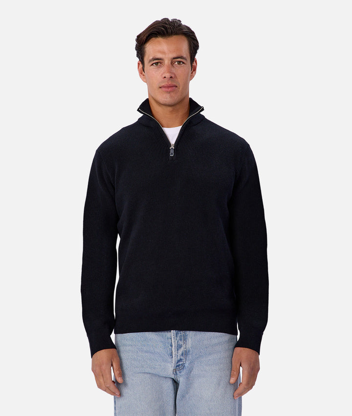 Man wearing a black quarter-zip sweater and light blue jeans on a white background