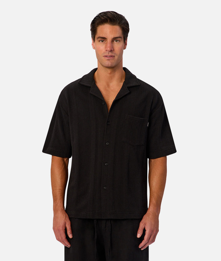 A man wearing a black short-sleeve button-up camp shirt with a chest pocket, photographed against a light gray studio background.
