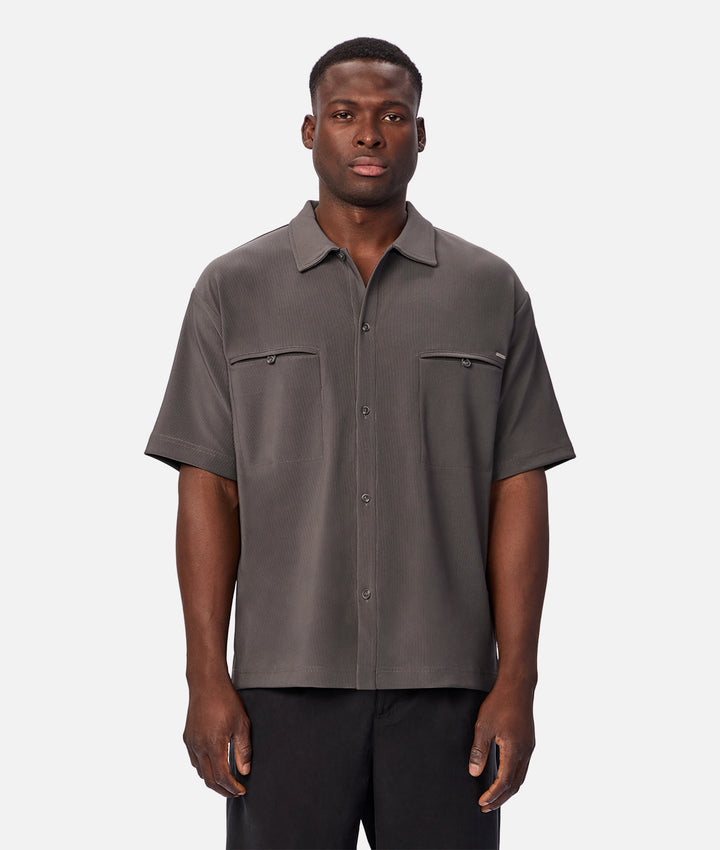A man wearing a short-sleeve button-up shirt in dark gray with two chest pockets, paired with black pants, standing against a light gray studio background.