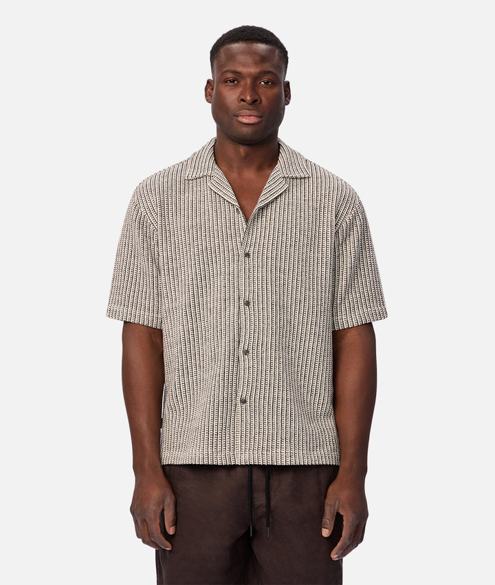 A man wearing a short-sleeve button-up shirt with a beige and black vertical textured stripe pattern, paired with dark brown drawstring pants, against a light gray background.