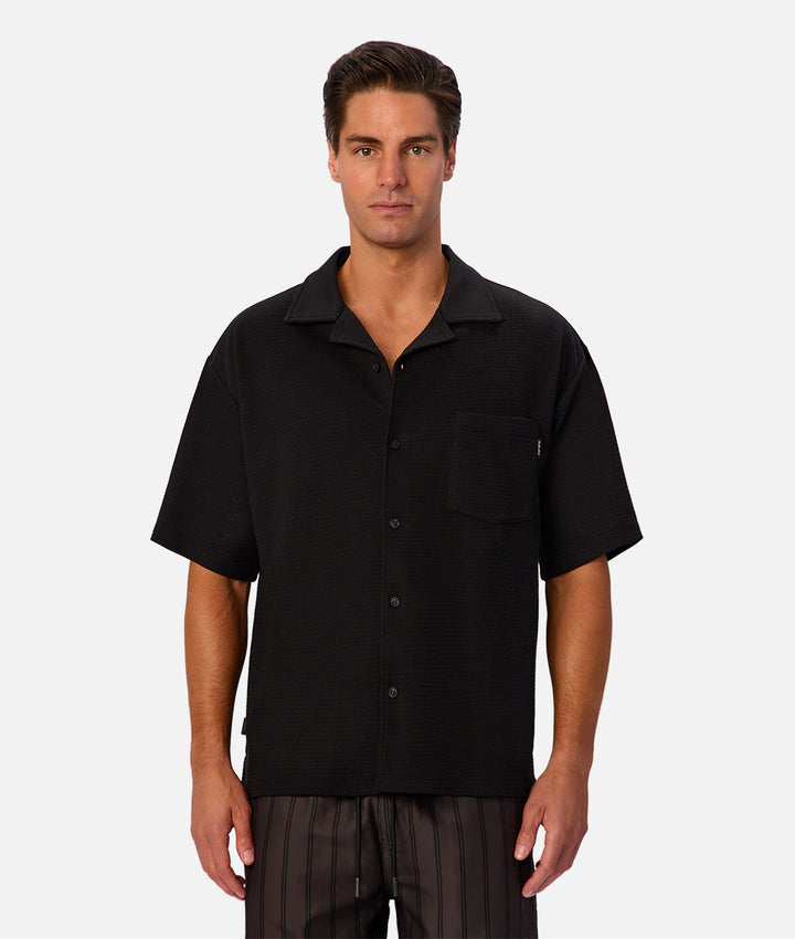 A man wearing a black short-sleeve button-up camp shirt with a chest pocket, paired with dark striped pants, photographed against a light gray studio background.
