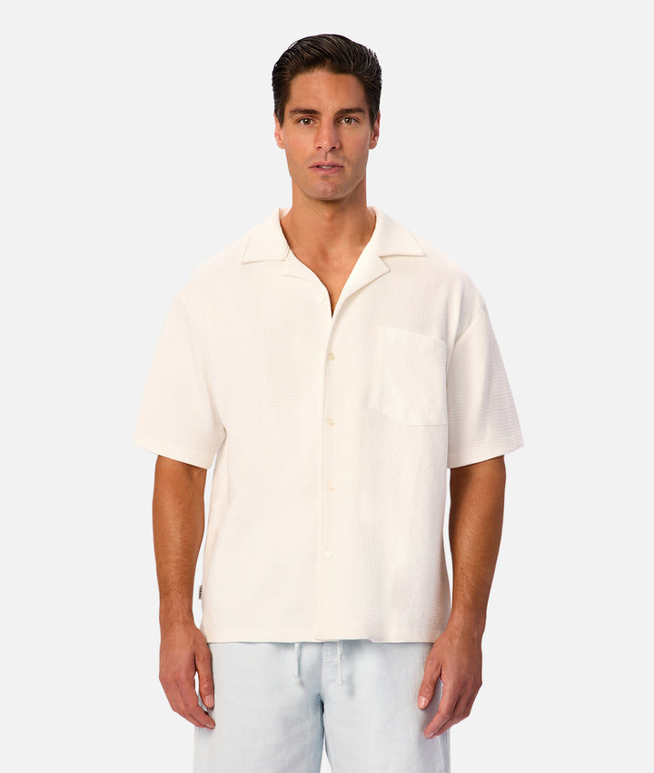 A man modeling a short-sleeve camp collar button-up shirt in off-white, paired with light blue pants, against a solid light gray background.