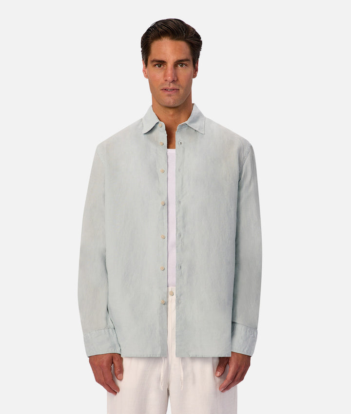 A man modeling a light sage green linen button-up shirt over a white undershirt and white trousers, set against a plain light gray background.