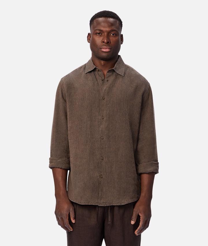 A man wearing a brown linen button-up shirt with rolled-up sleeves and dark brown pants, standing against a light gray studio background.