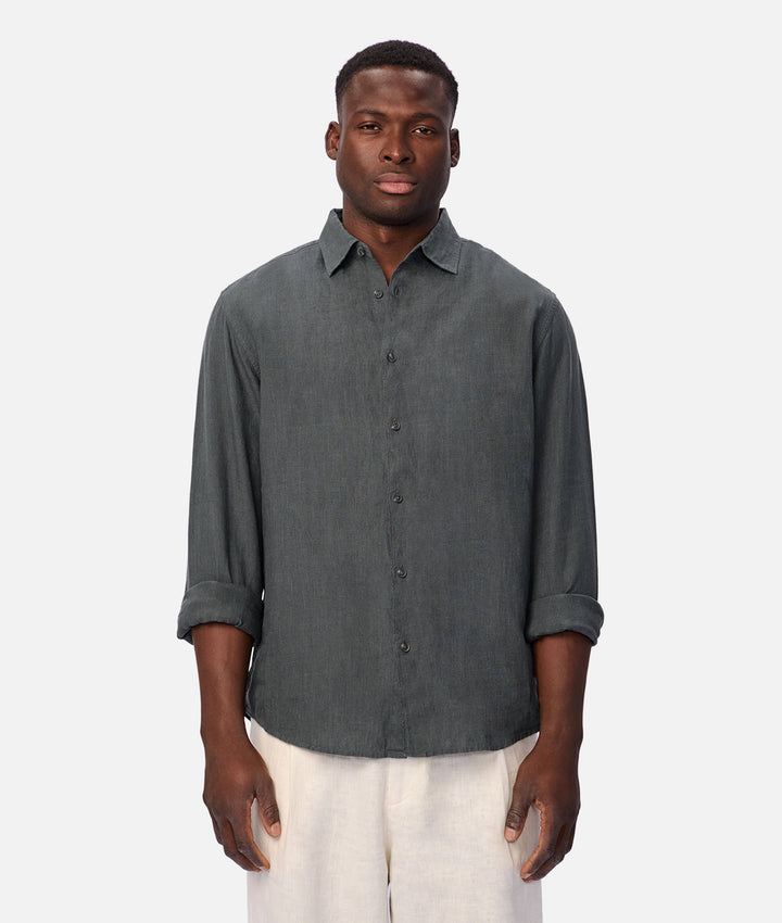 A man wearing a dark gray linen button-up shirt with rolled-up sleeves and off-white trousers, standing against a light gray background.