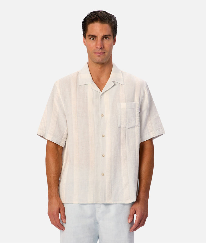 A man wearing a short-sleeve button-up camp shirt in a light cream or off-white color with subtle vertical stripes, paired with light blue jeans, against a solid light gray background.