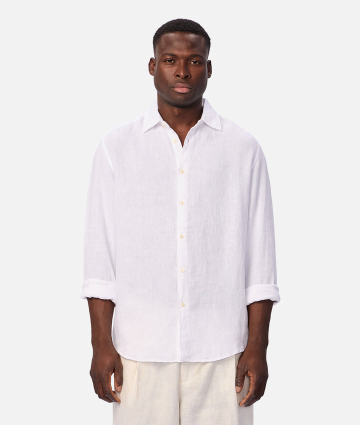 A man wearing a white long-sleeve linen button-up shirt with rolled-up sleeves and light beige trousers, standing against a solid light gray background.