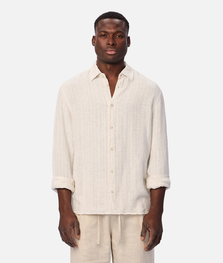 A man wearing a light beige linen button-up shirt with rolled-up sleeves and matching beige drawstring pants, standing against a solid light gray background.