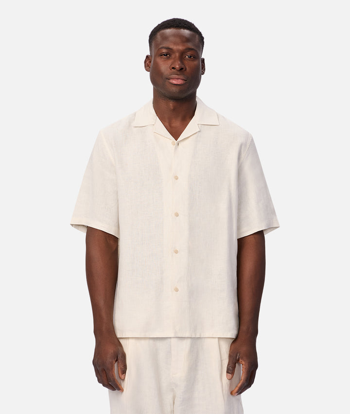 A man wearing a short-sleeve button-up shirt and matching shorts in a light cream linen fabric, photographed against a solid light gray studio background.