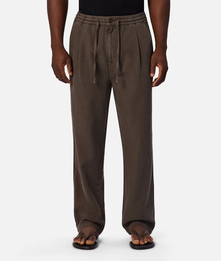 A person wearing loose, wide-leg drawstring pants in a dark brownish-gray color, paired with a black top and brown sandals, shown against a plain light gray studio background.