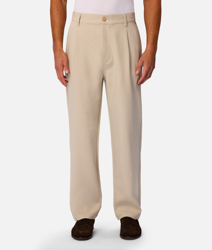 A person wearing light beige pleated trousers with a gold button closure, paired with dark brown suede loafers, shown against a plain light gray studio background.