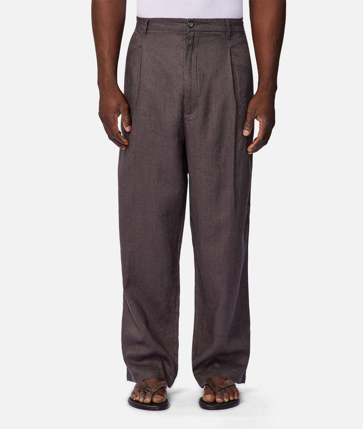 A person wearing loose, wide-leg charcoal gray trousers with a white top and brown sandals, shown from the waist down against a light gray studio background.