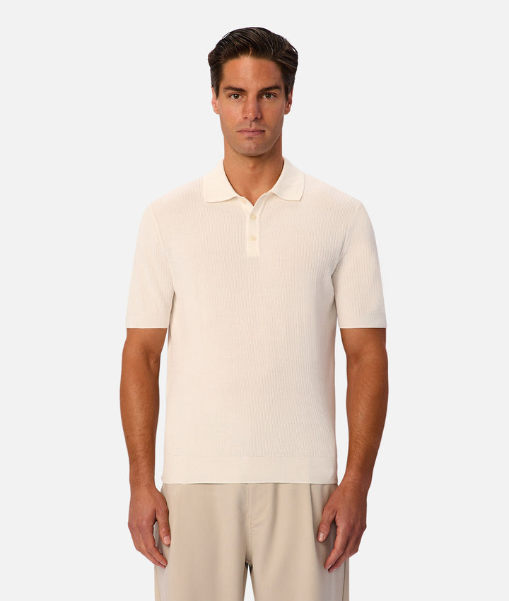 A man modeling a short-sleeve ribbed knit polo shirt in cream, paired with light beige trousers, against a solid light gray background.