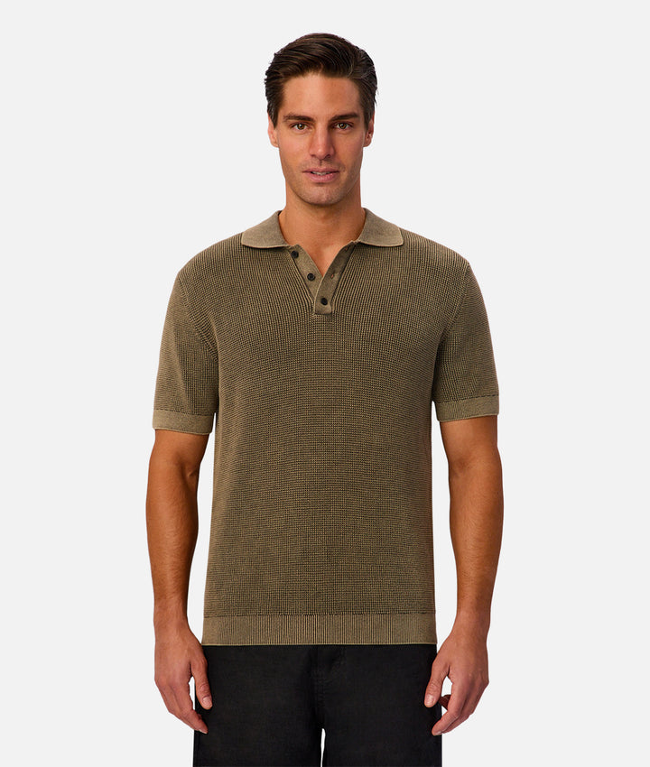A man wearing a short-sleeve knit polo shirt in olive green with a three-button placket, standing against a light gray studio background.