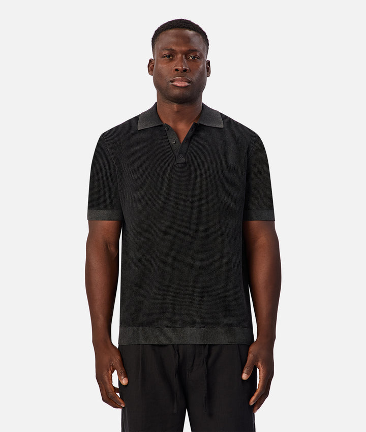 A man wearing a dark charcoal gray ribbed knit polo shirt with a darker gray collar, cuffs, and hem, paired with black trousers, against a light gray background.