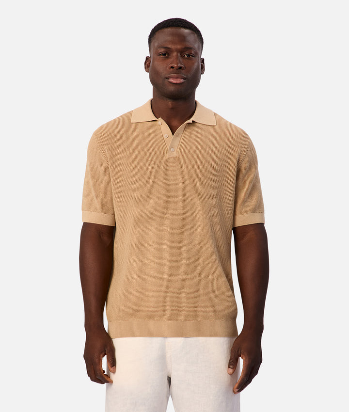 A man wearing a beige short-sleeve knit polo shirt with a matching collar and button placket, paired with white trousers, against a light gray studio background.