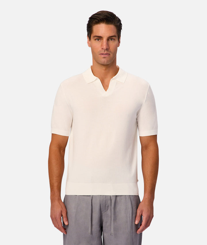 A man modeling a short-sleeve knit polo shirt in off-white, paired with gray drawstring pants, against a light gray studio background.