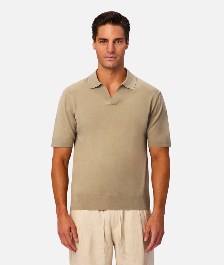 A man wearing a short-sleeve knit polo shirt in a light taupe-beige color, paired with light beige trousers, standing against a solid light gray background.