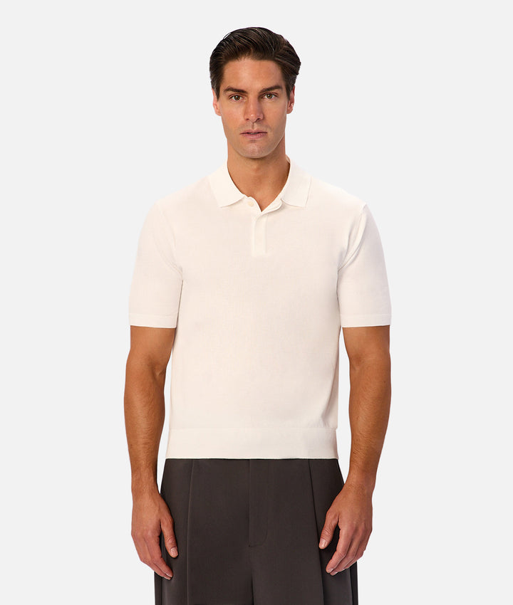 A man wearing a short-sleeve knit polo shirt in off-white, paired with dark gray trousers, standing against a light gray studio background.