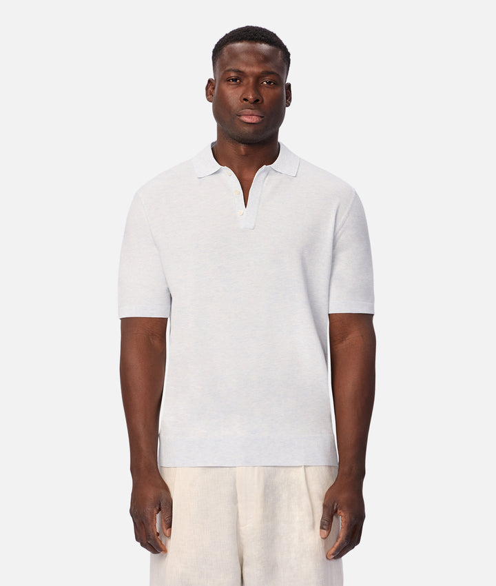 A man wearing a light gray short-sleeve polo shirt and off-white trousers, standing against a solid light gray background.