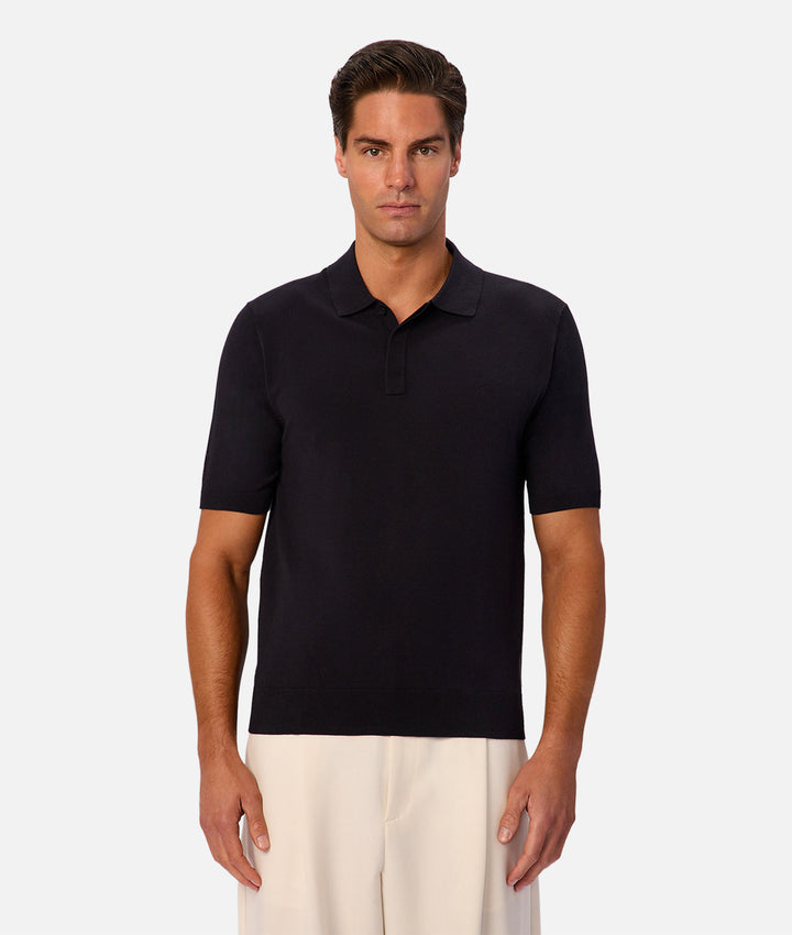A man wearing a short-sleeve knit polo shirt in solid black, paired with off-white trousers, standing against a light gray studio background.